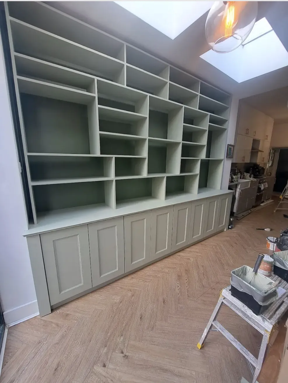 Full-wall fitted shelving in sage green with an asymmetric grid of shelves above panelled cupboards